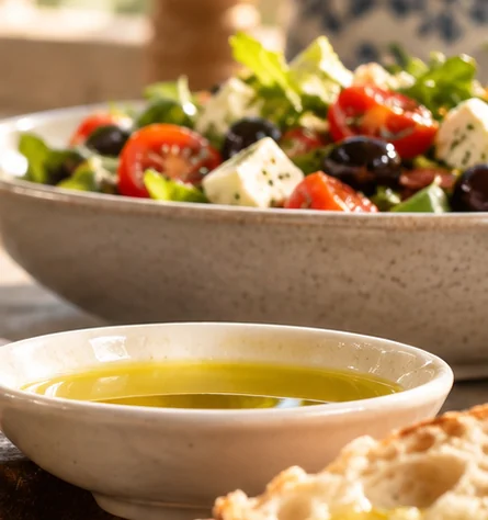 Coupelle d'huile d'olive dorée à côté d'une salade de tomates, olives et fromage, et de pain frais
