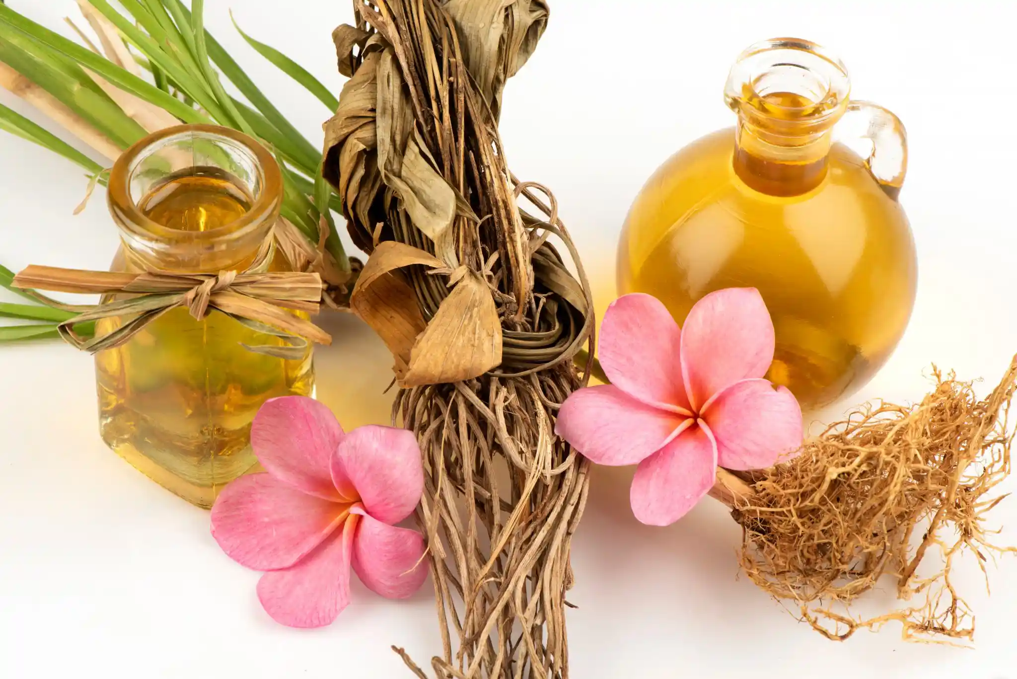 Bouteilles d’huile de vétiver entourées de racines séchées et de fleurs tropicales roses, sur fond blanc.