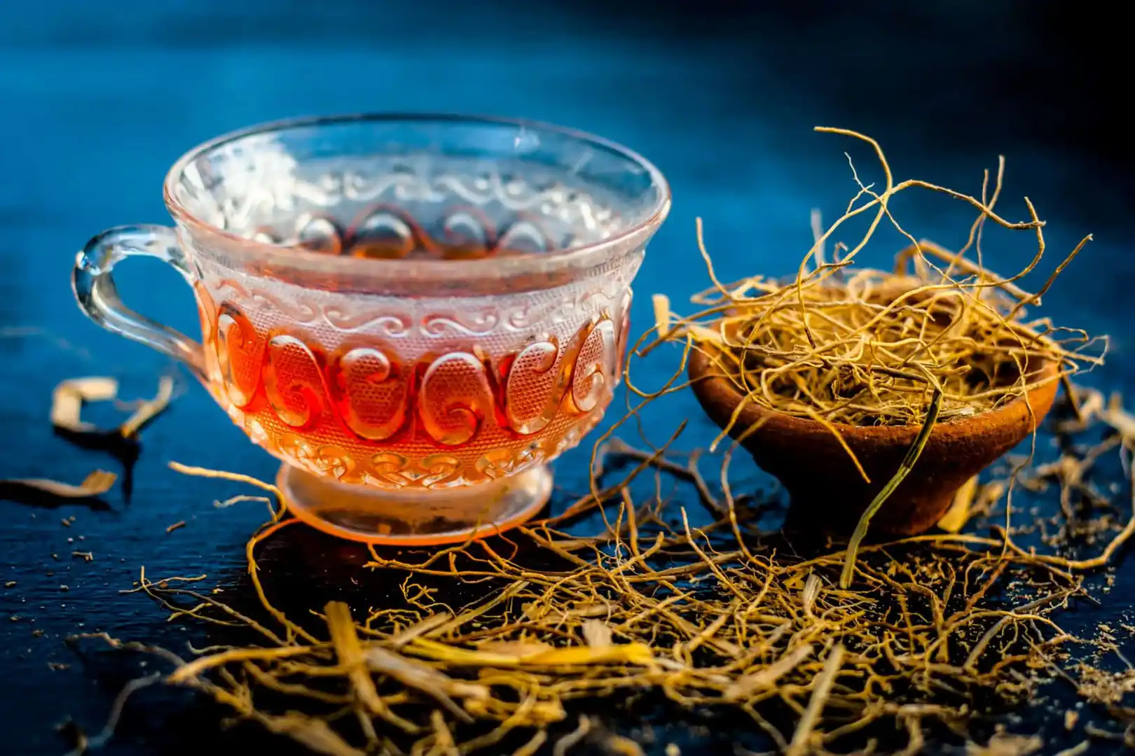 Tasse de tisane rougeâtre à base de racine de vétiver, posée à côté d’un bol contenant les racines séchées de la plante.