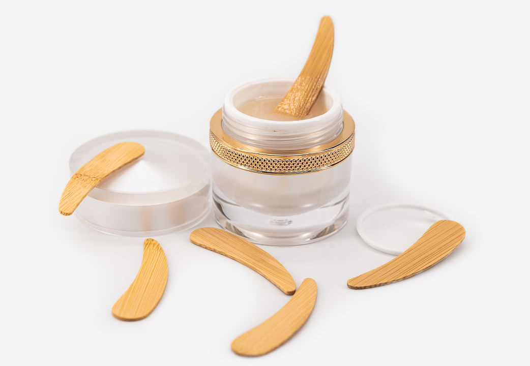 Pot en verre ouvert contenant du musc tahara blanc crémeux avec applicateur en bambou, entouré de spatules en bambou sur fond blanc