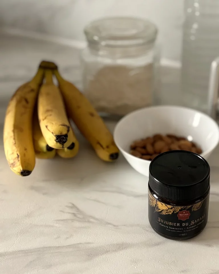 Bananes mûres, bocal de flocons d’avoine, bol d’amandes et pot de miel de jujubier du Sahara Natural Fawaid sur un plan de travail en marbre