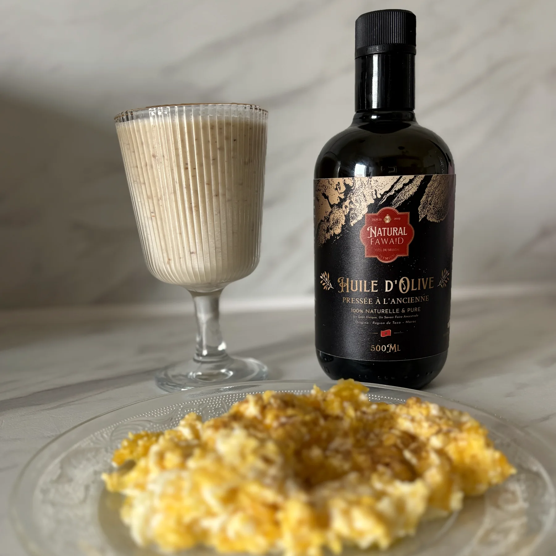 Verre de smoothie banane dattes aux amandes à côté d'une bouteille d'huile d'olive Natural Fawaid et d'une assiette d'omelette au cumin